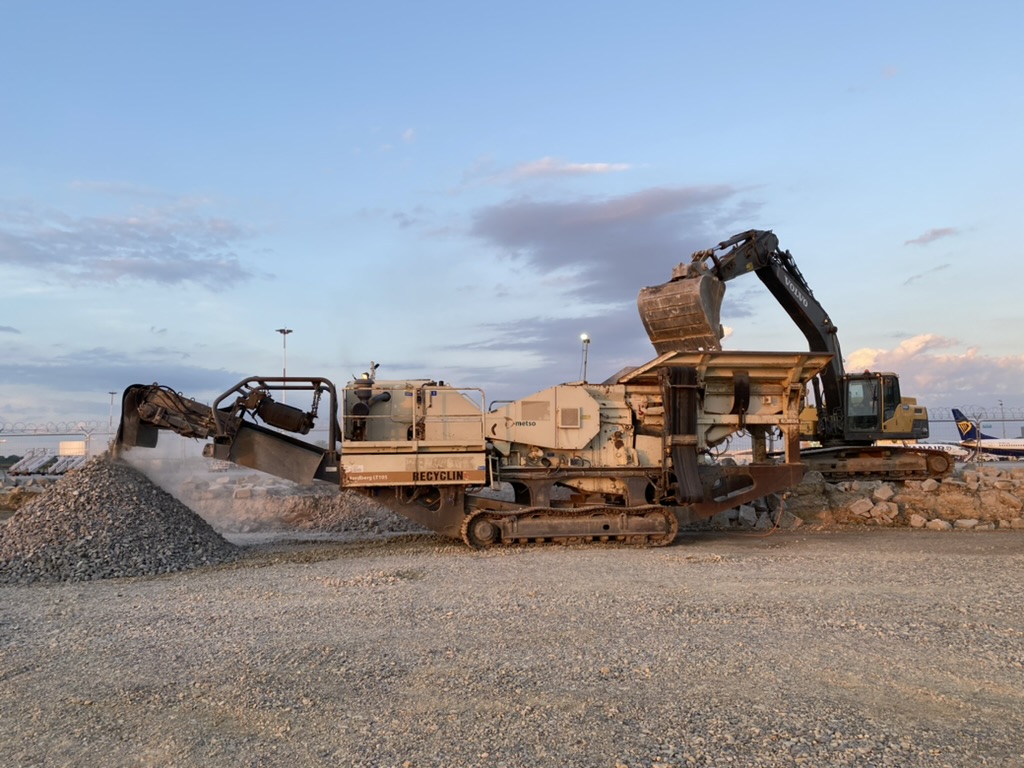 Heavy machinery sorting and piling gravel at a construction site under a partly cloudy sky.
