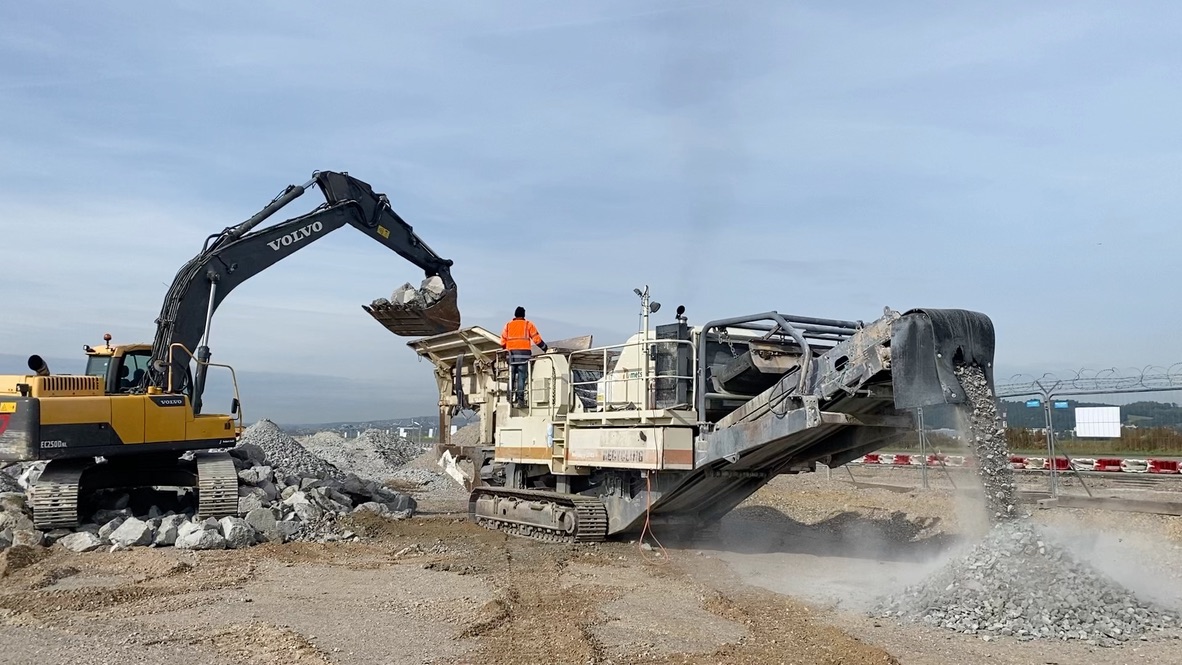 Yellow Volvo excavator loading rocks into machine that sorts gravel on construction site.