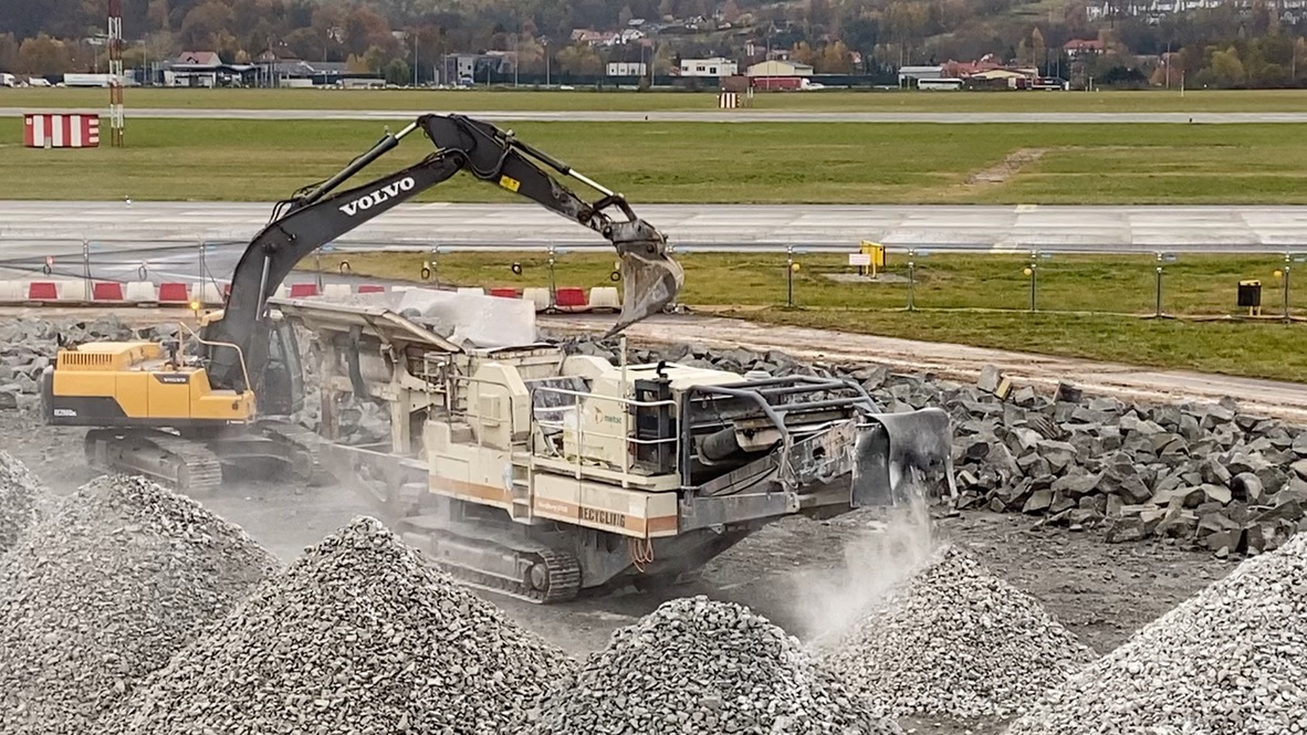 Excavator loading rocks into crusher machine, with piles of crushed gravel nearby in an open field.