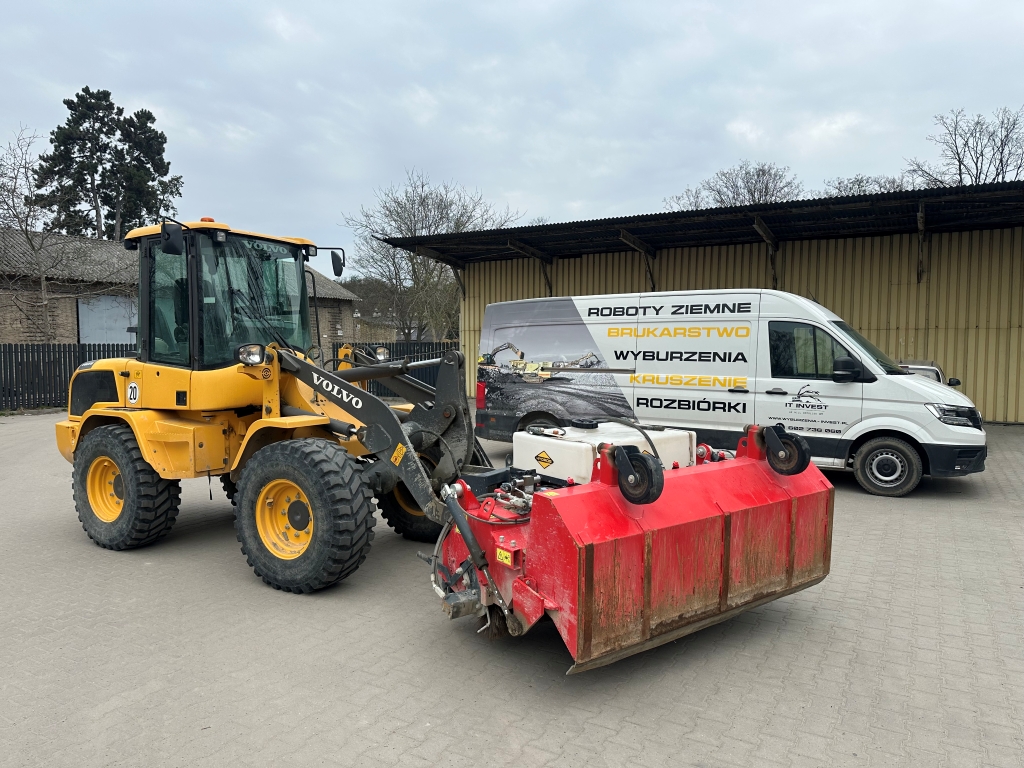 Yellow Volvo construction loader parked near a white van with Polish text and a red attachment.