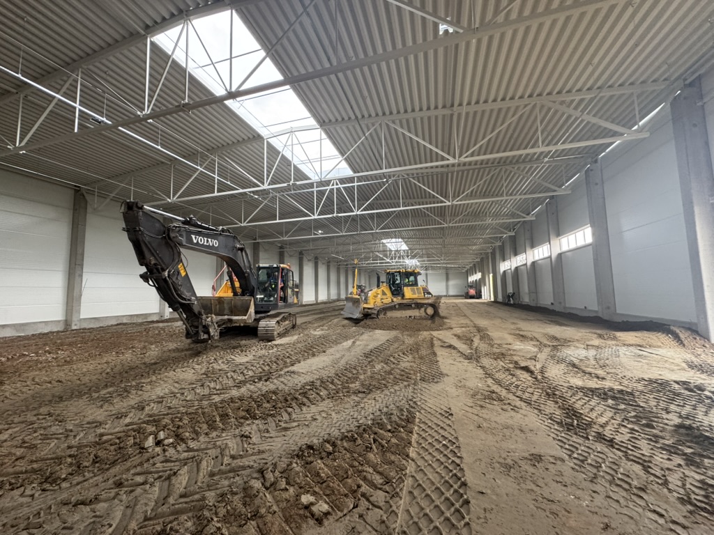 Interior of large industrial building with excavator and bulldozer on dirt floor under metal roof.