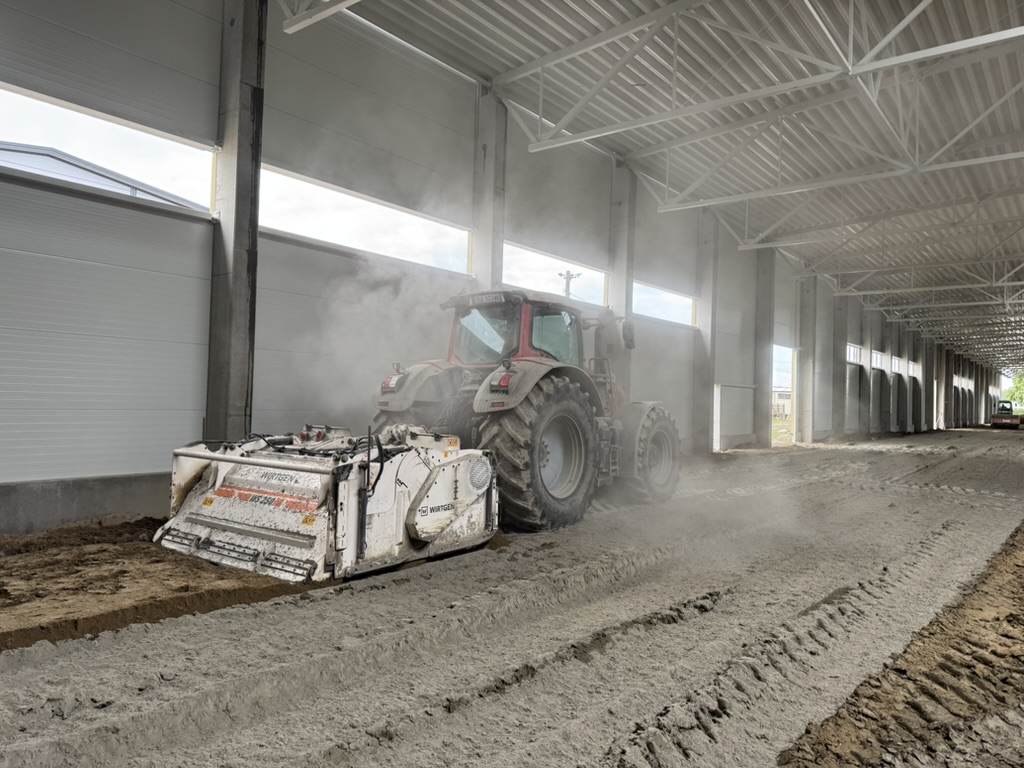Heavy machinery smoothing dirt floor inside a large industrial building under construction.