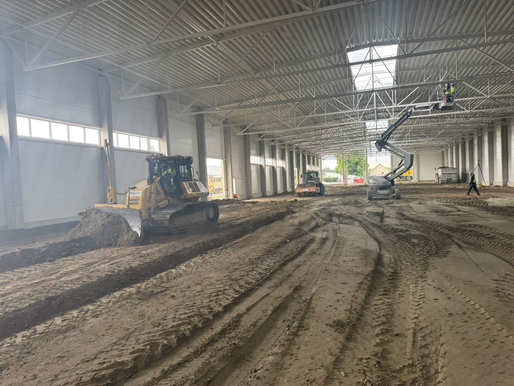 Indoor construction site with bulldozer, workers on lift, dirt floor, and construction equipment.
