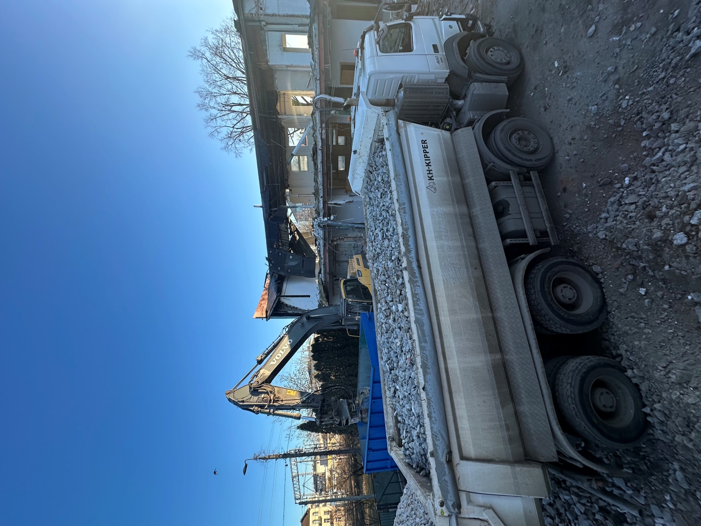 Construction site with a white KH-KIPPER truck loaded with gravel and a Volvo excavator loading debris.