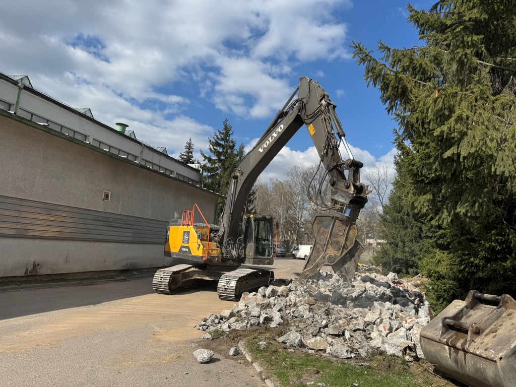 Excavator breaking concrete rubble near a building and pine trees under a cloudy blue sky.
