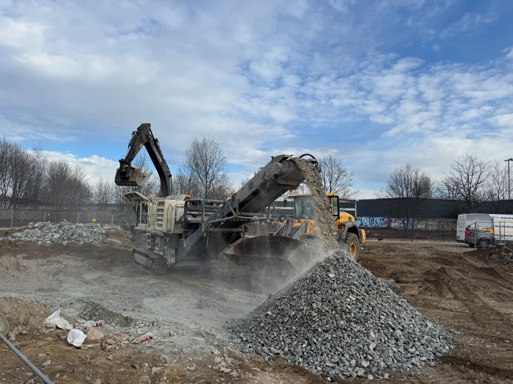 Construction site with machinery moving and piling crushed rocks under a cloudy sky.