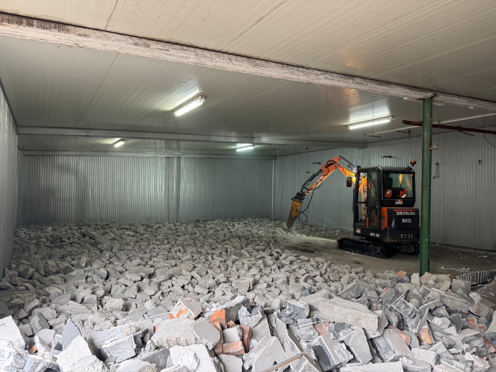 Indoor demolition with excavator breaking concrete floor covered in rubble under fluorescent lights.