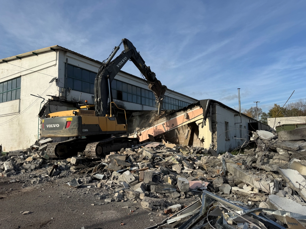 Yellow Volvo excavator demolishing a concrete building with rubble scattered around.