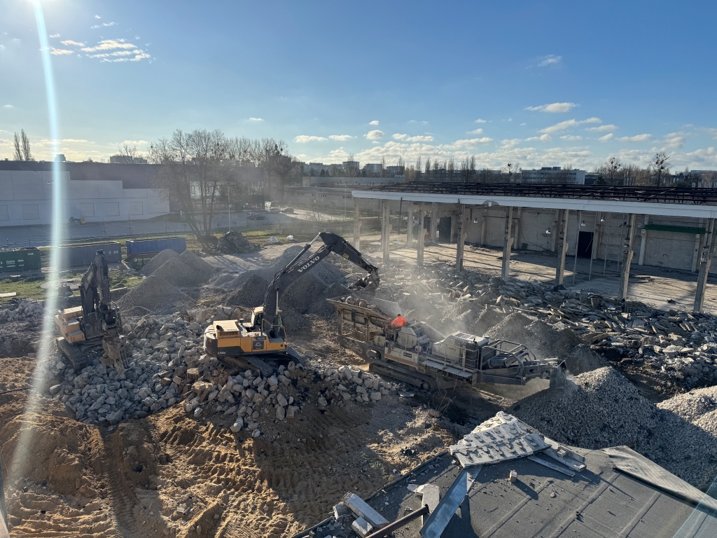 Construction site with excavators moving rocks and debris near a partially demolished building under blue sky.