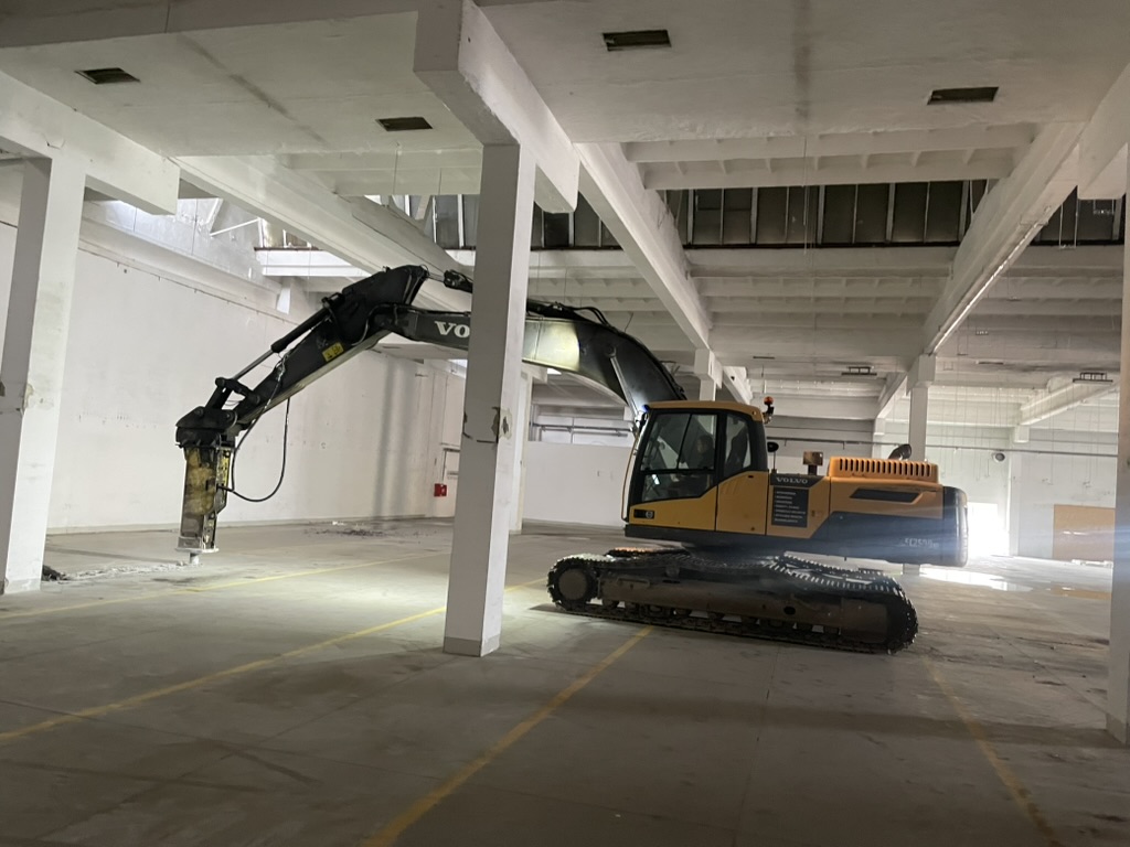 Yellow Volvo excavator with hydraulic breaker inside a large empty industrial building.