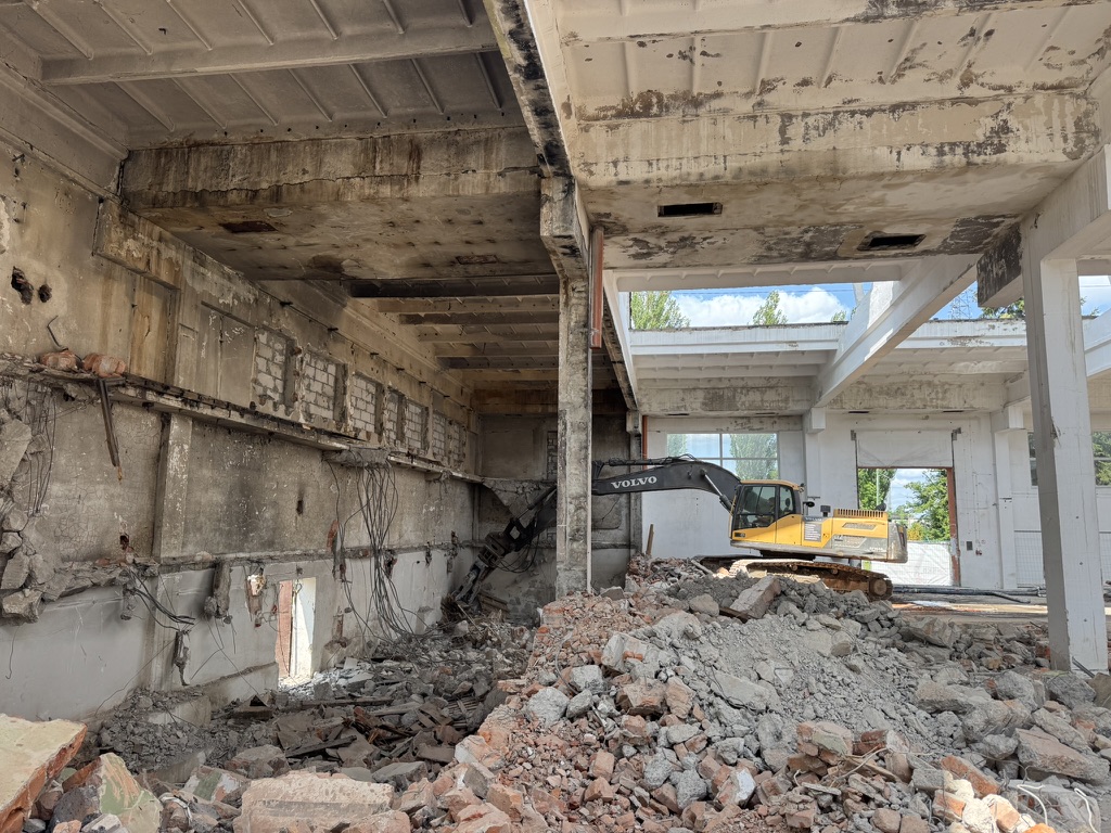 Yellow Volvo excavator inside a partially demolished building with rubble piled on the floor.