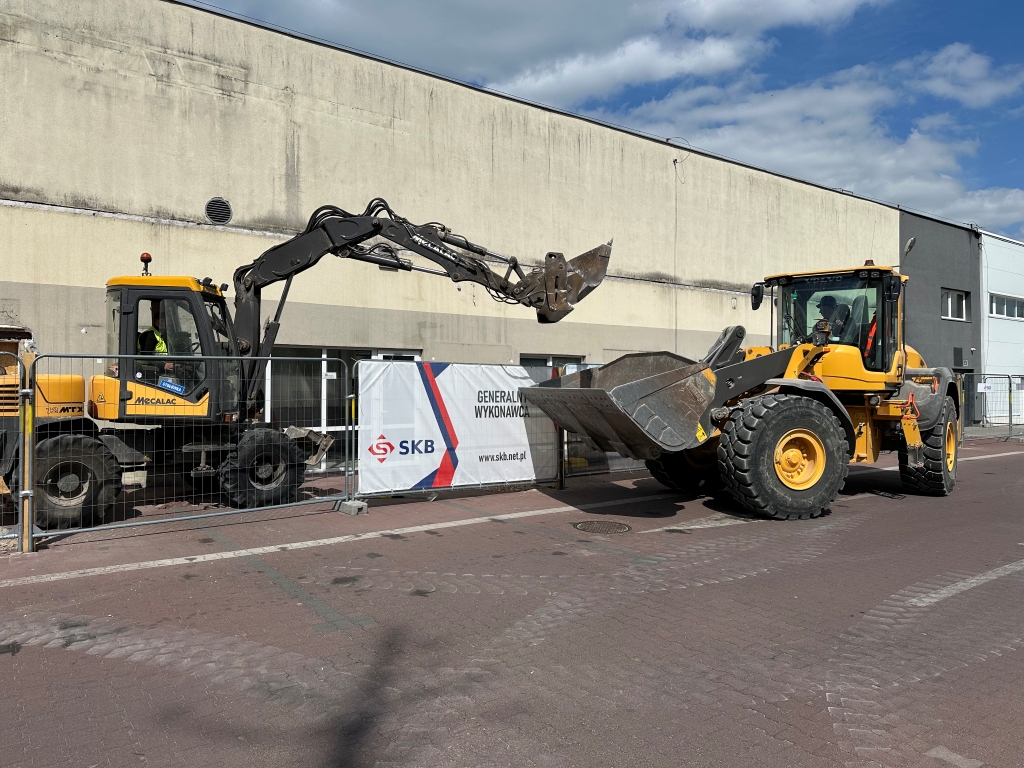 Two yellow construction machines behind a metal fence on a paved street near a beige building.