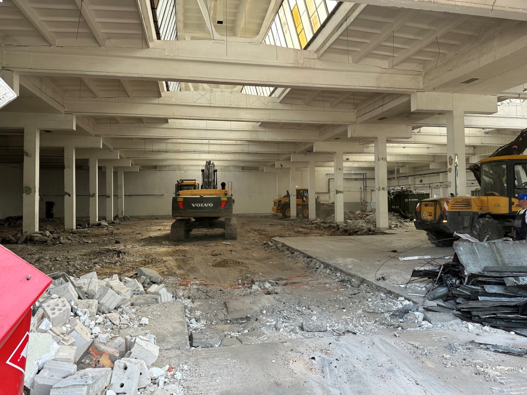 Interior of a large warehouse under renovation with construction machinery and rubble on the ground.