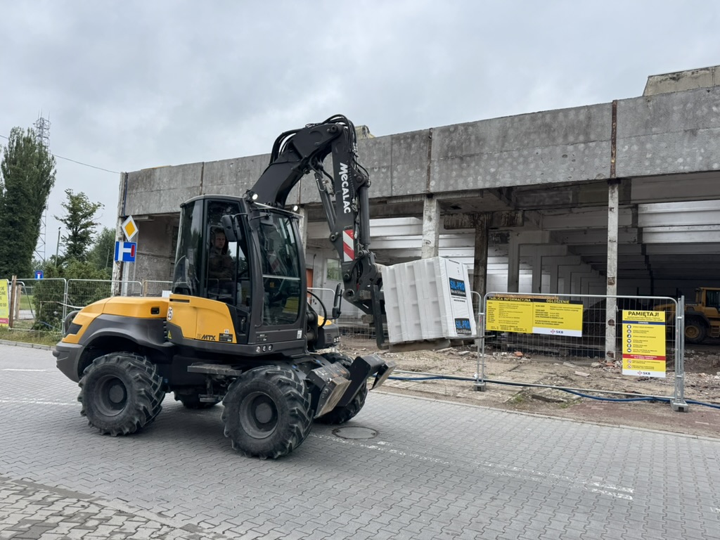 Yellow and black Mecalac excavator lifting a pallet in front of a concrete construction site.