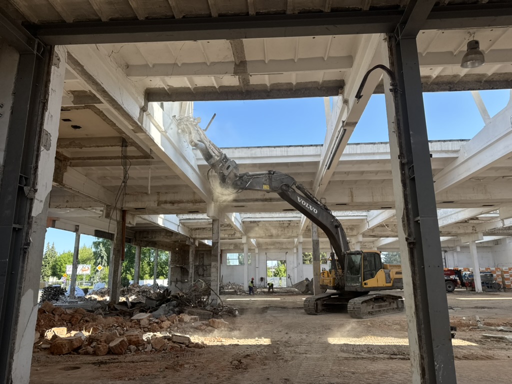 Excavator demolishing concrete ceiling inside worn industrial building with debris on the ground.