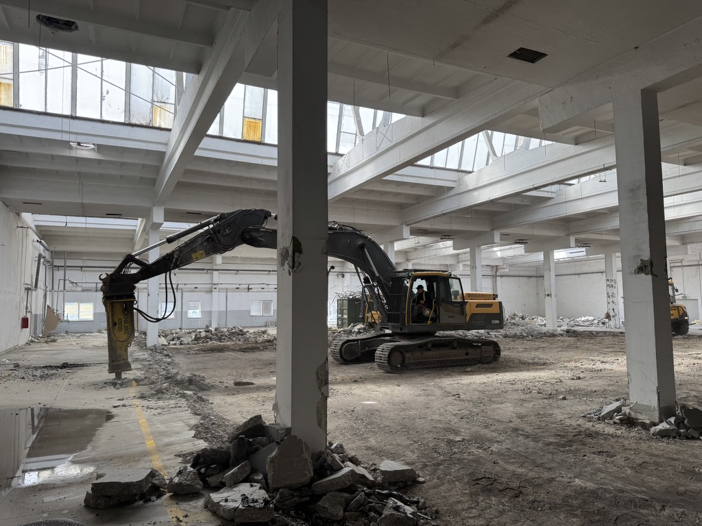 Excavator machine inside a large warehouse with broken concrete and rubble on the ground.