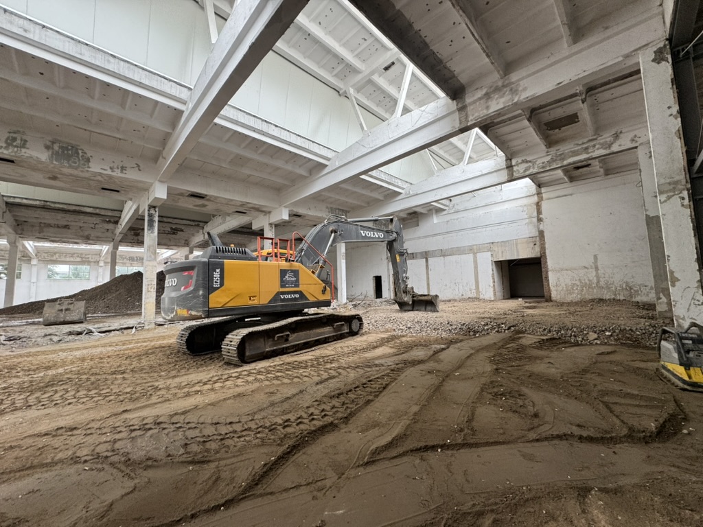 Yellow and black Volvo excavator inside a large, empty, white-walled industrial building with dirt floor.