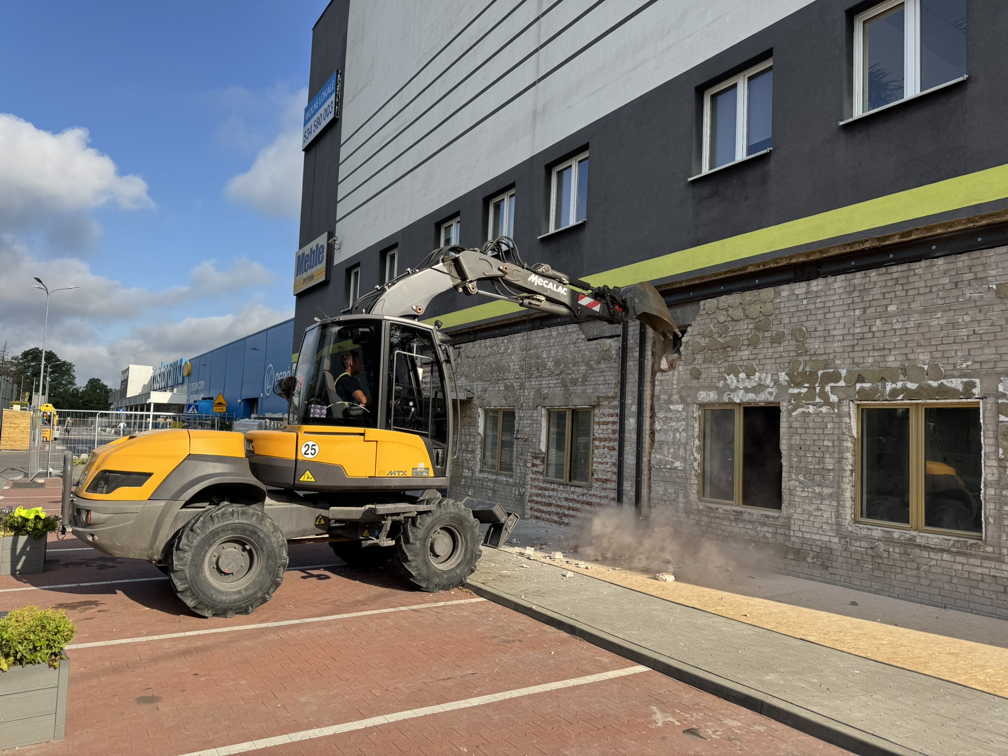Yellow excavator demolishes brick wall of gray building, creating dust on sunny day.