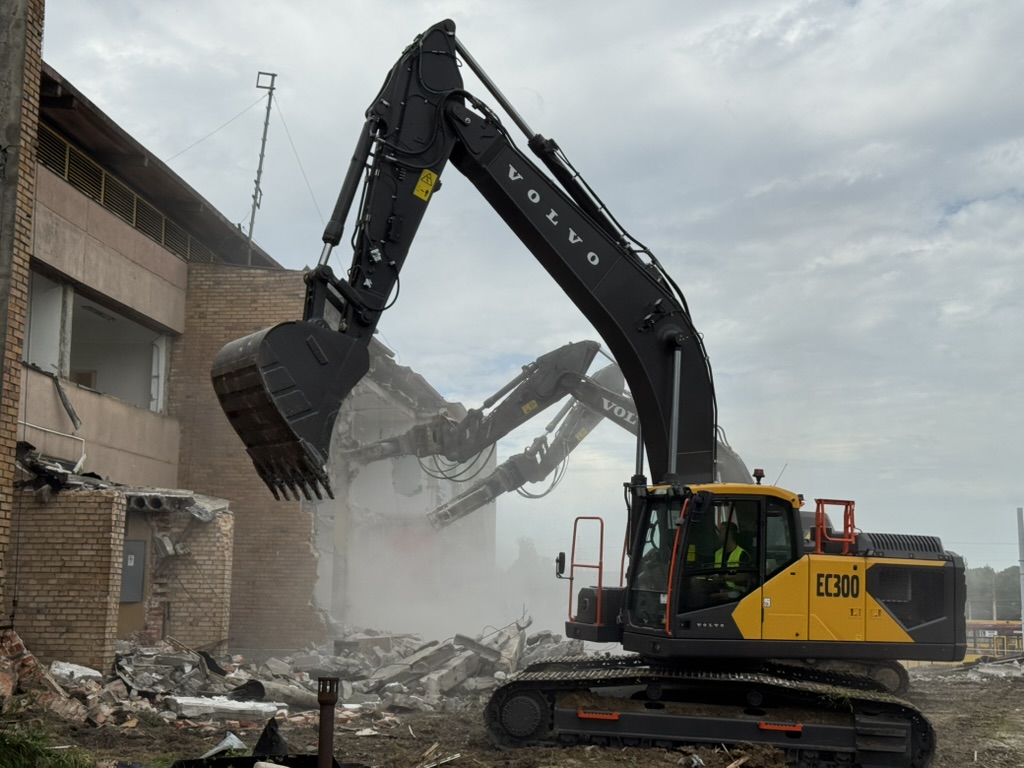 Yellow and black Volvo EC300 excavator demolishing a brick building with rubble on the ground.