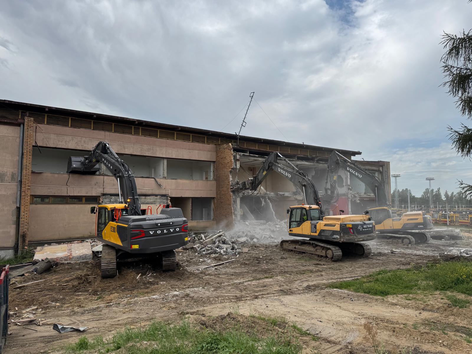 Three Volvo excavators demolishing a two-story building amid rubble under a cloudy sky.