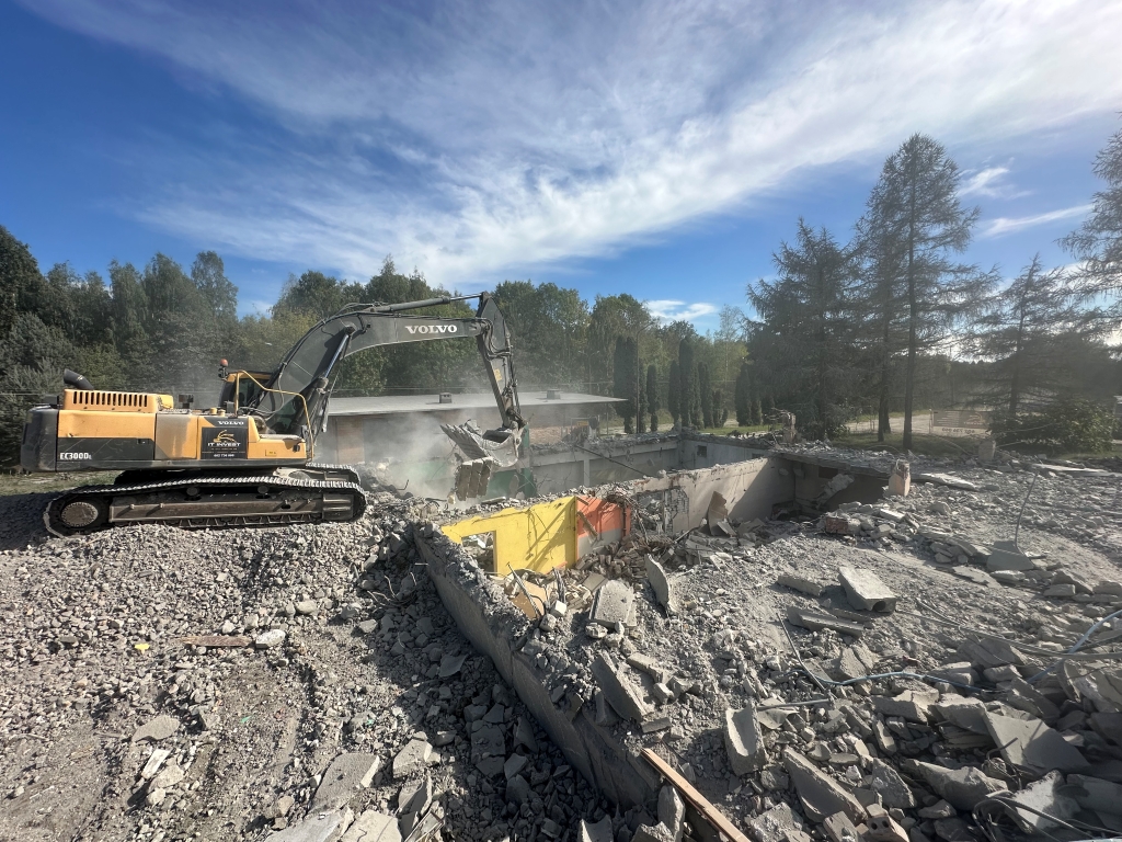 Yellow Volvo excavator demolishing a concrete building amid rubble under a blue sky with scattered clouds.