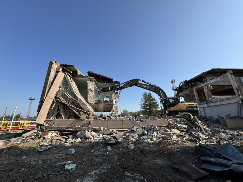 Excavator demolishing a partially destroyed building with rubble scattered around on a clear day.
