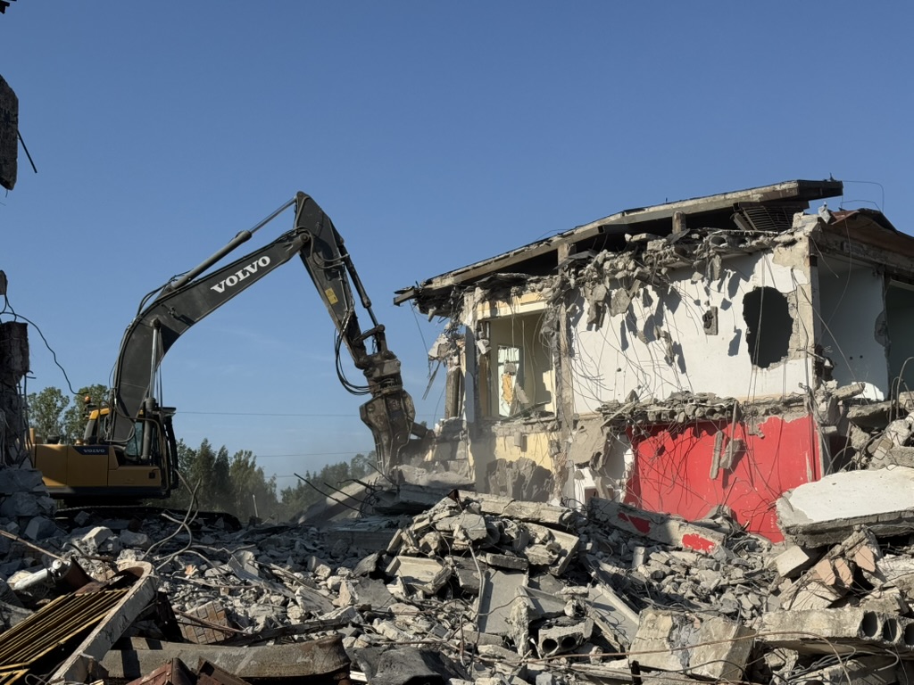 Volvo excavator demolishing a partially destroyed concrete building under clear blue sky.