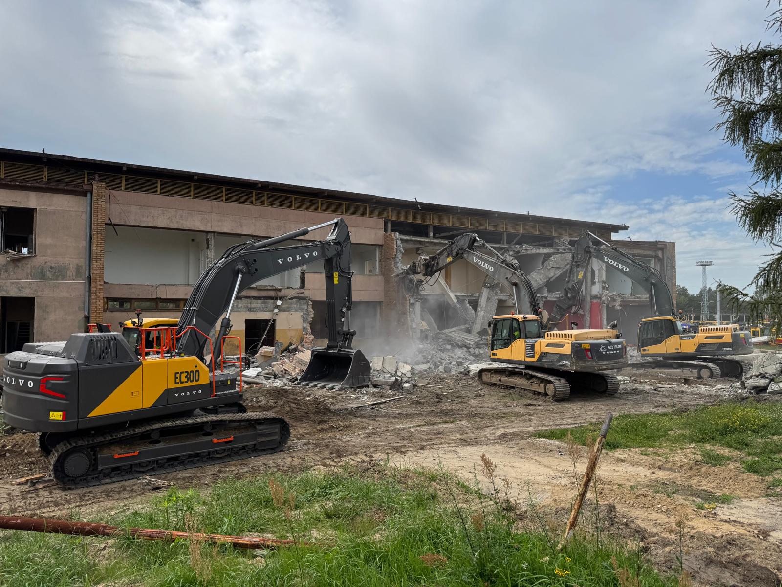Three yellow Volvo excavators demolishing a concrete building under cloudy sky.
