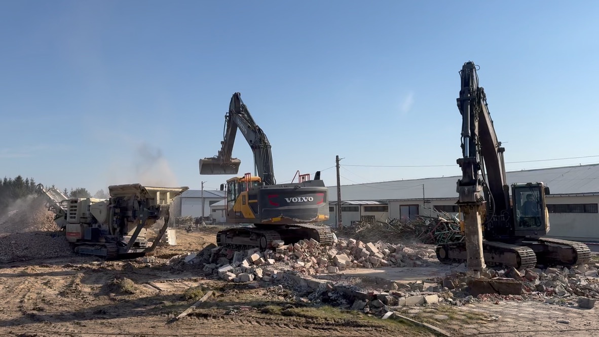 Two excavators and a stone crusher working on rubble at a demolition site near warehouse buildings.