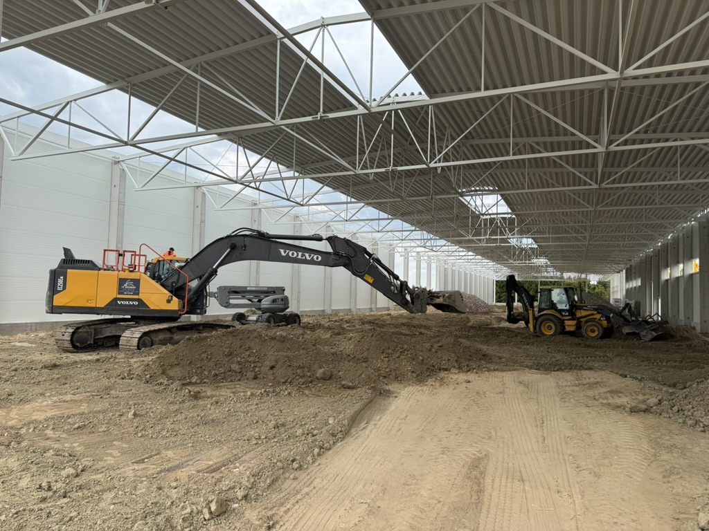Inside a large industrial building, two excavators move and level dirt on the ground.