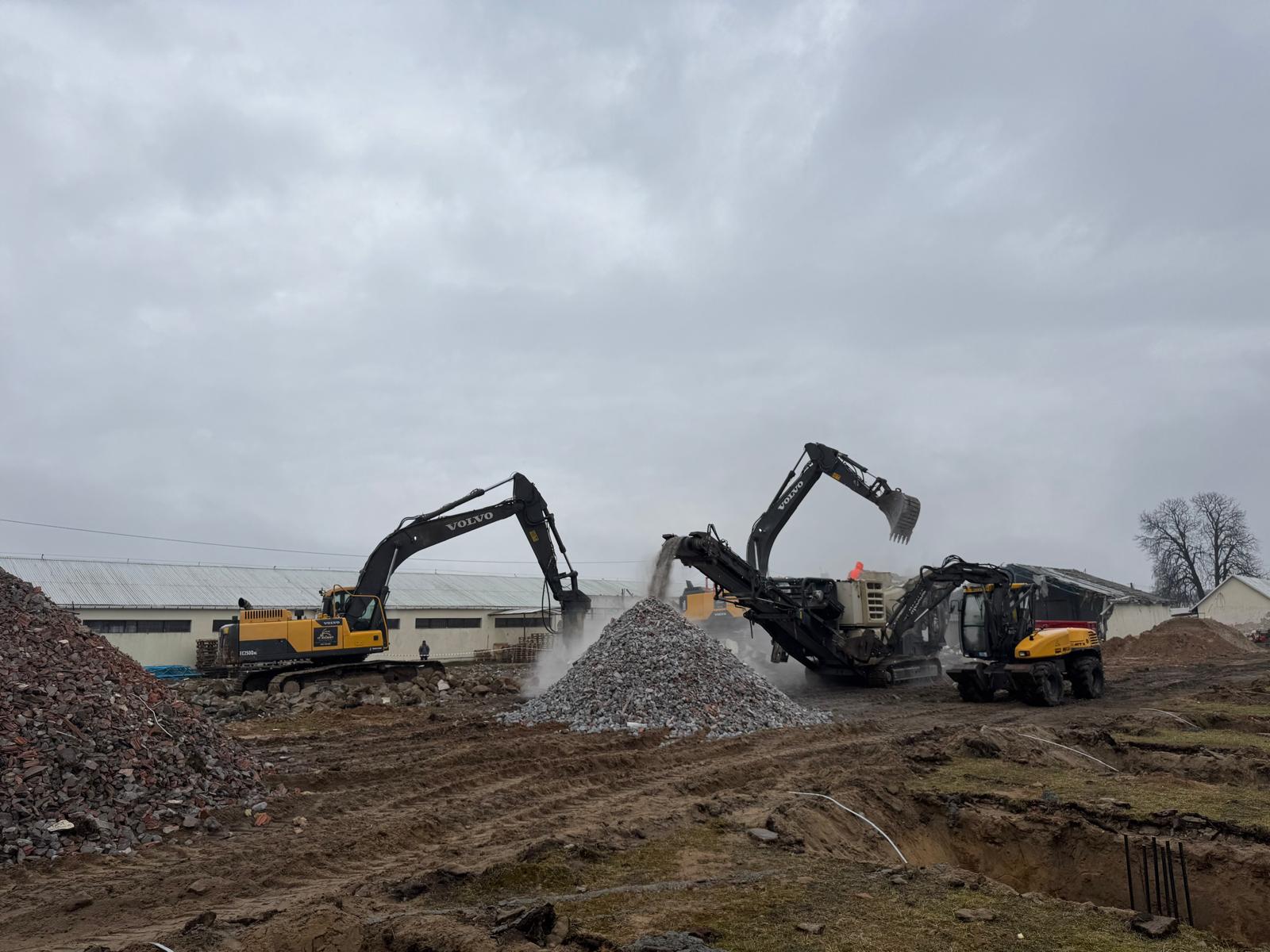 Construction site with three Volvo excavators moving and piling rubble under a cloudy gray sky.
