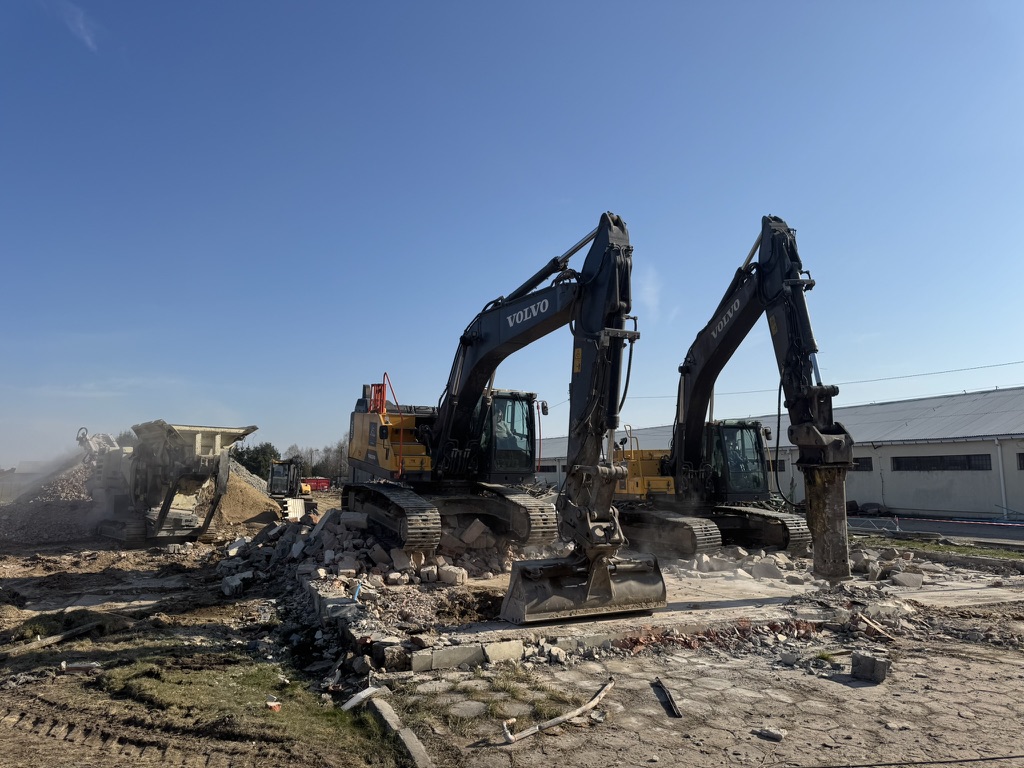 Two Volvo excavators demolishing a concrete and brick surface at a construction site under clear blue sky.