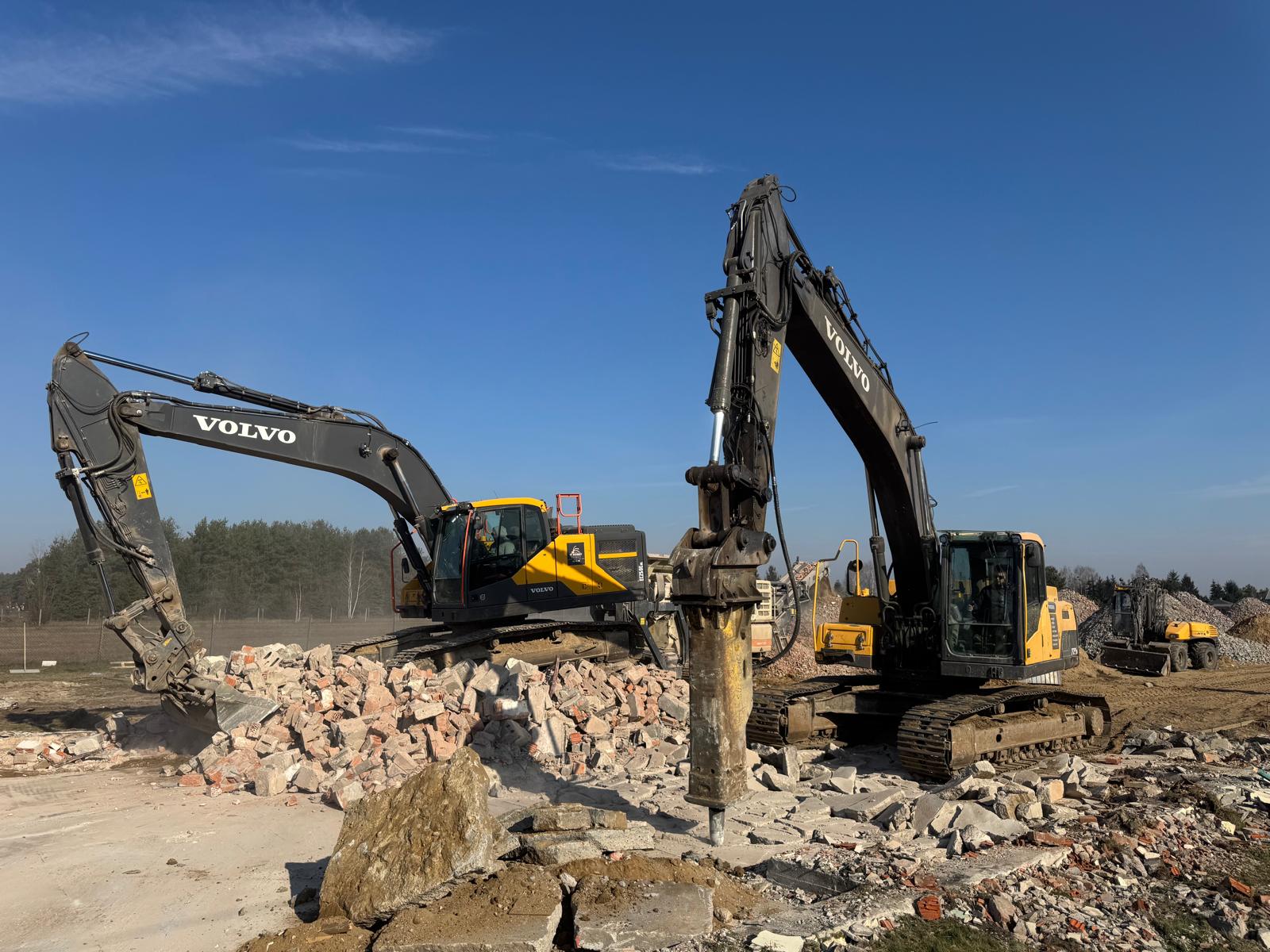 Two Volvo excavators working among rubble under a clear blue sky on a construction site.