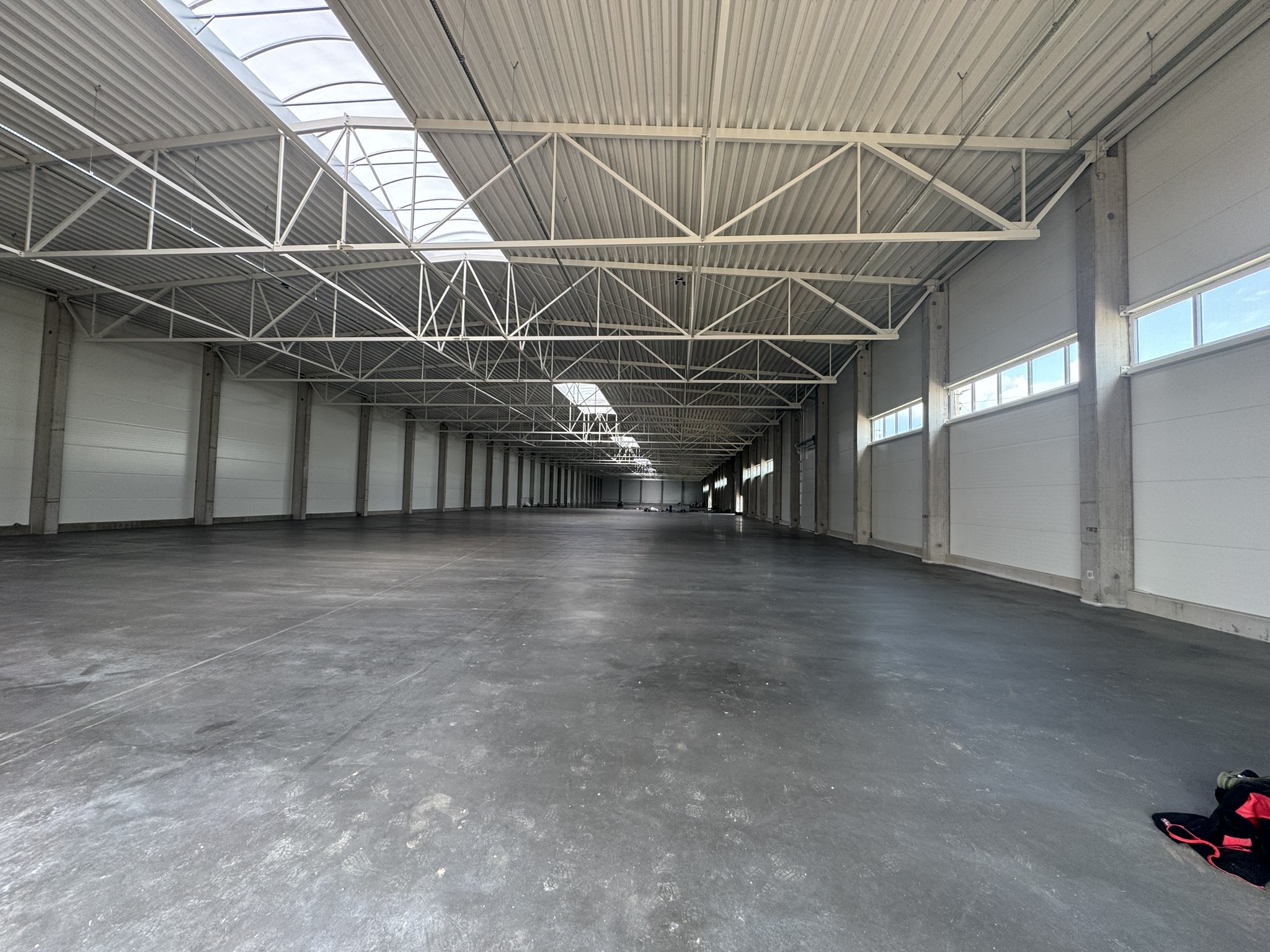 Large empty warehouse with concrete floor, white walls, skylights, and metal ceiling trusses.