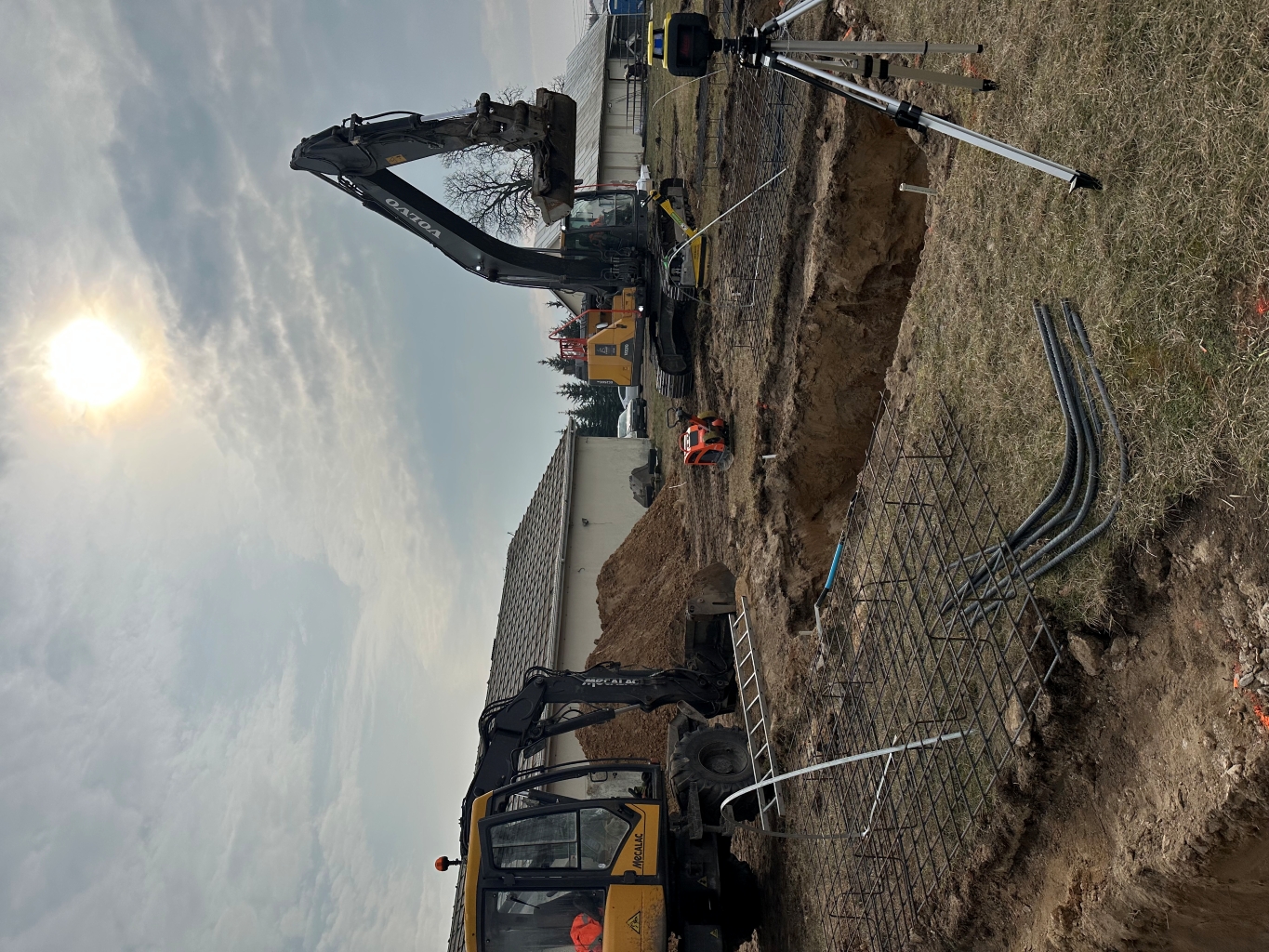 Construction site with excavators, metal rebar grids, a tripod with surveying equipment, and a partly cloudy sun.
