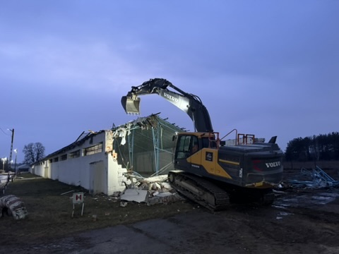 Excavator demolishing a building at dusk with rubble on the ground nearby.