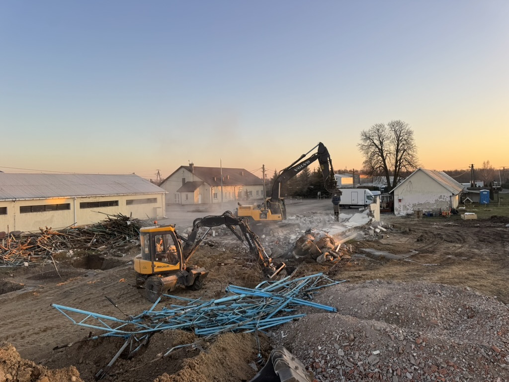 Two excavators demolish a building at a construction site during sunset with debris and houses in background.