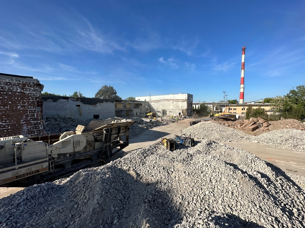 Construction site with rubble piles, heavy machinery, and a tall red and white smokestack under a blue sky.