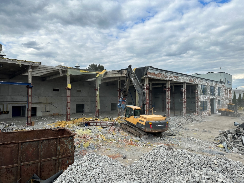 Excavator demolishing an old industrial building surrounded by rubble under a cloudy sky.