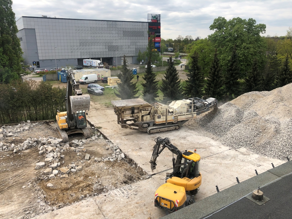Construction site with two excavators and a rock crusher near rubble piles and a modern industrial building.
