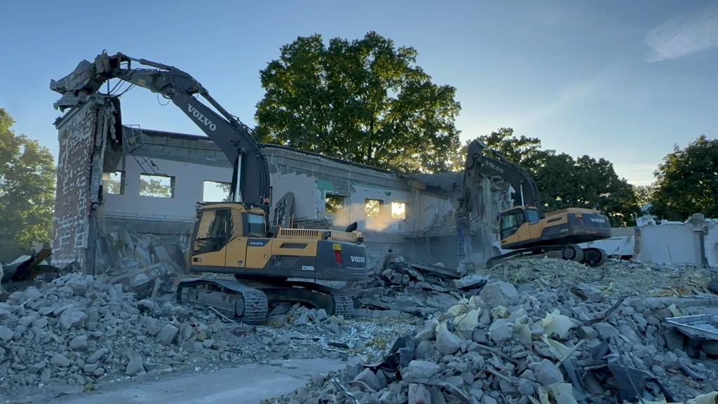 Two Volvo excavators demolishing a large building surrounded by rubble and trees in the background.