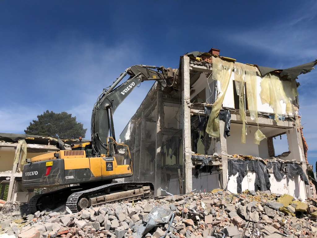 Yellow Volvo excavator demolishing a partially destroyed multi-story building with debris piled in front.