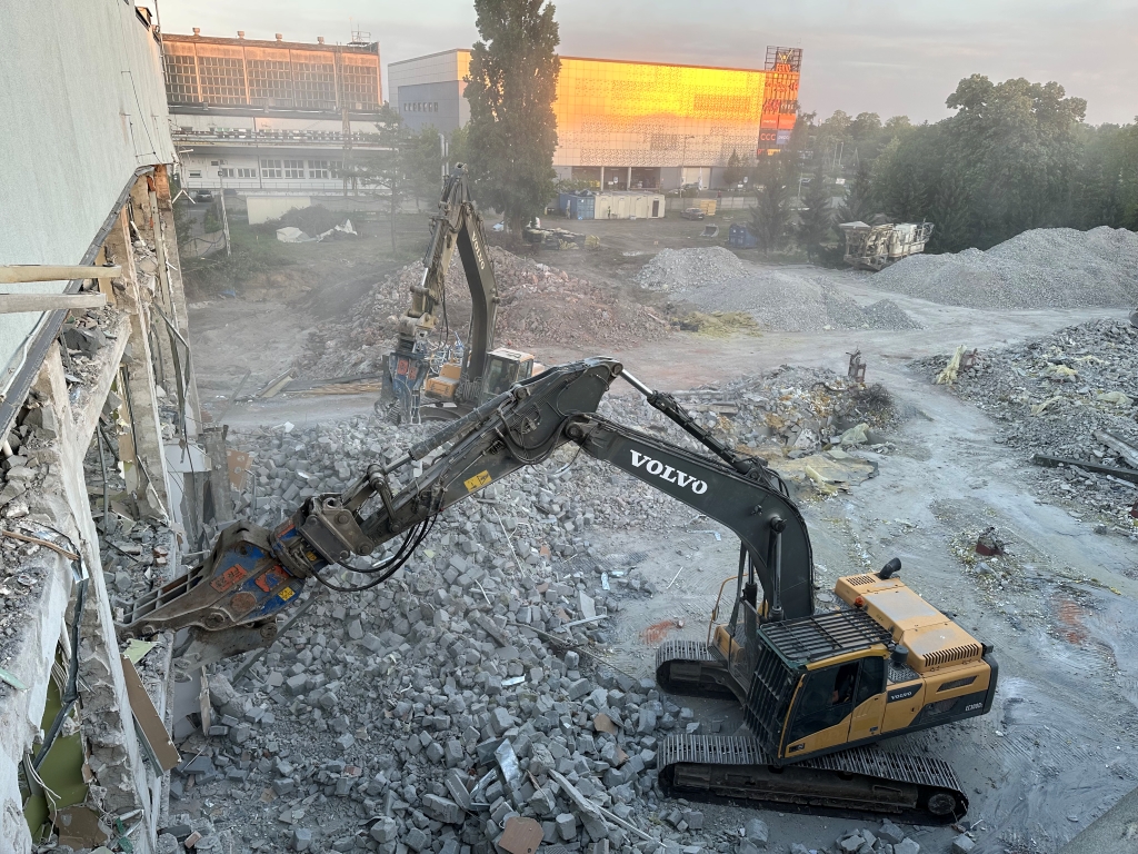Volvo excavator demolishing a concrete building amid rubble and construction debris at a worksite.