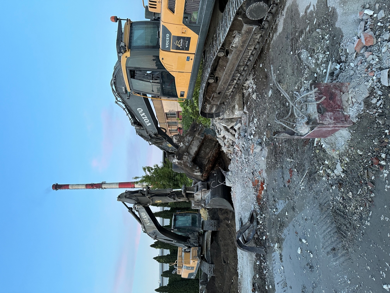 Two Volvo excavators working on a construction site with rubble and a tall smokestack in the background.