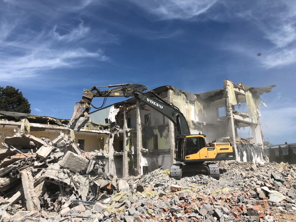 Yellow Volvo excavator demolishing a multi-story building amid rubble under a blue sky.