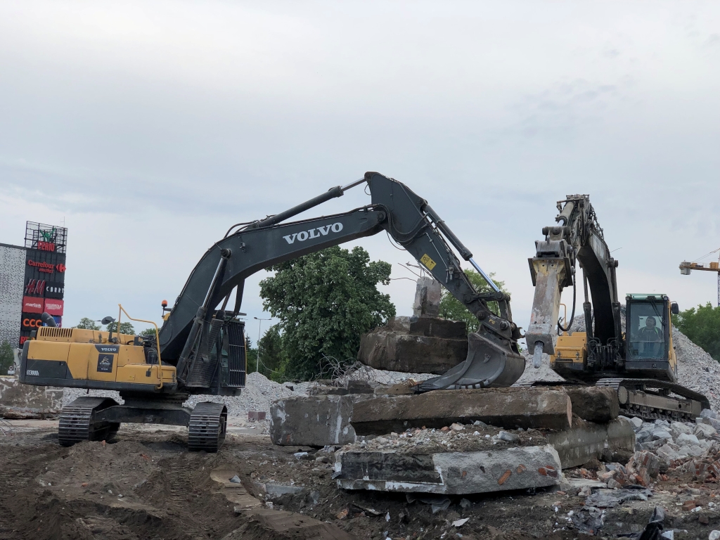 Two excavators moving large concrete slabs on a rubble-filled construction or demolition site.