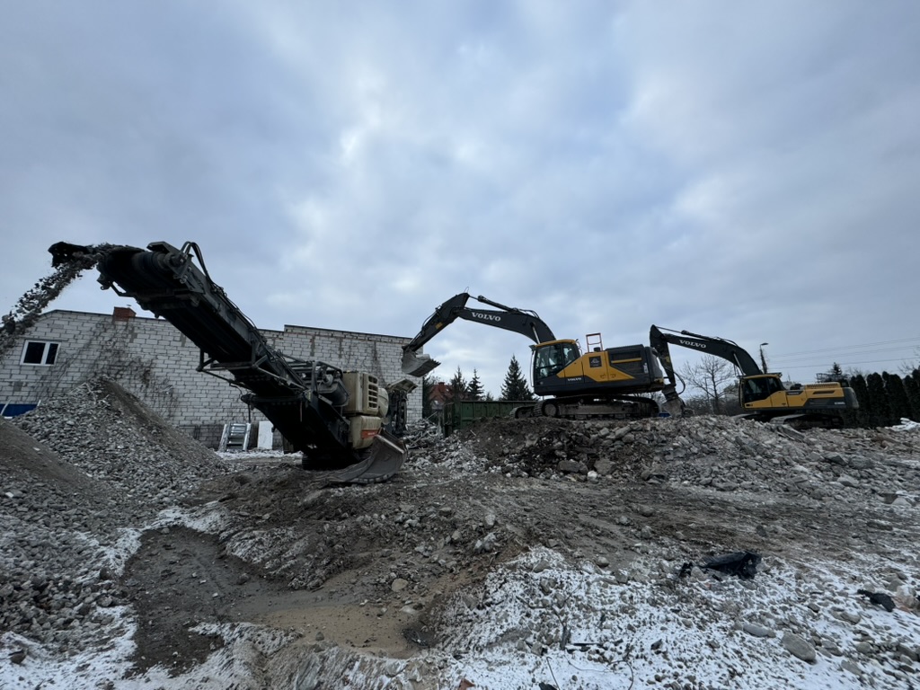 Two Volvo excavators move rubble on a construction site with a conveyor belt dumping debris on a pile.