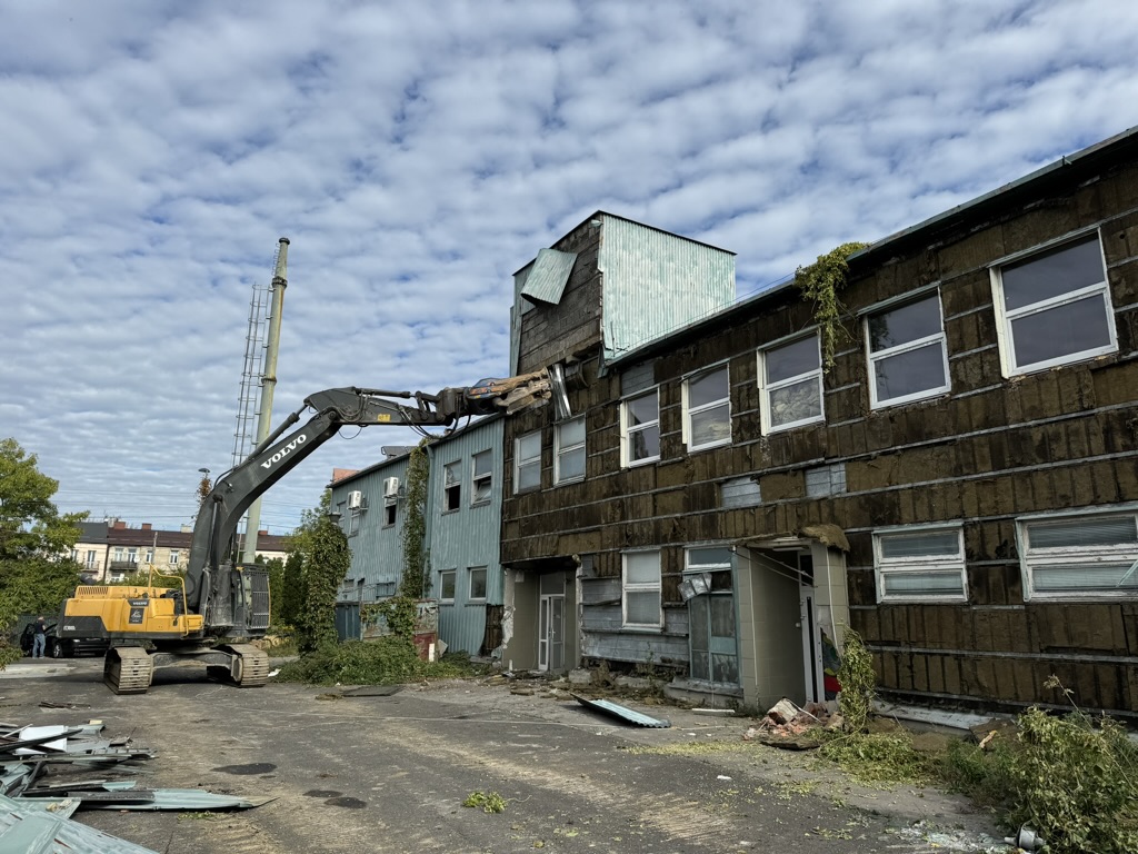 Excavator demolishes old blue industrial building under a cloudy sky with debris on ground.