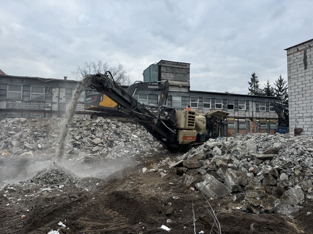 Excavator with conveyor sorting rubble amid demolished building remains under cloudy sky.