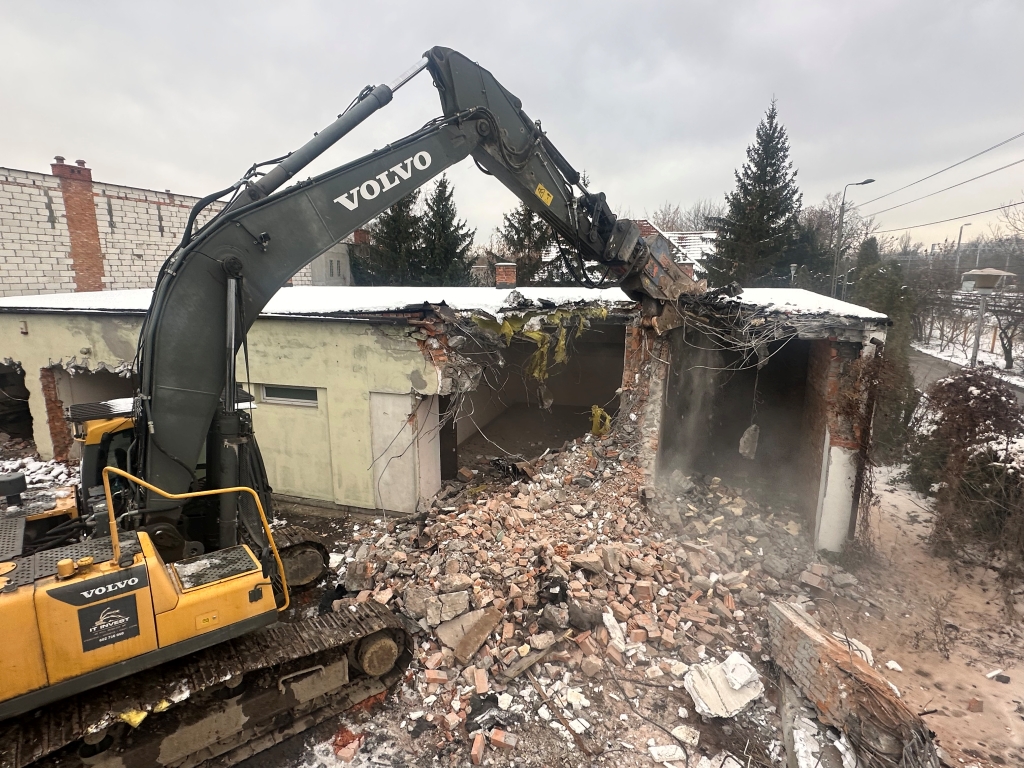 Yellow Volvo excavator demolishing a brick and concrete building amid snow and trees in winter.